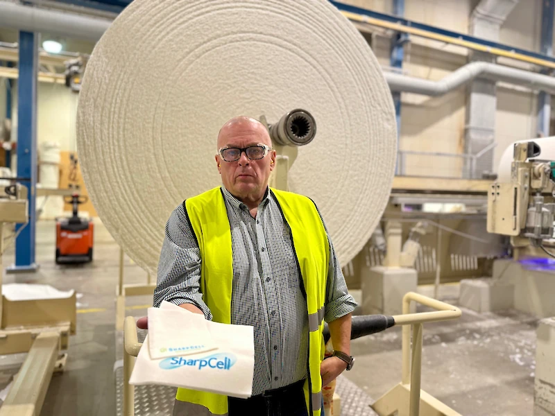 Seppo Saarisen taustalla paperikoneelta valmistunut konerulla, joka leikataan asiakkaan toiveiden mukaisiin kokoihin. Saarisen kädessä näkyy esimerkki valmiista tuotteesta.